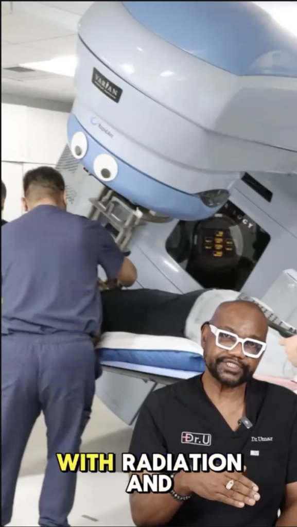 Dr. U in black scrubs speaking in front of a large radiation treatment machine.