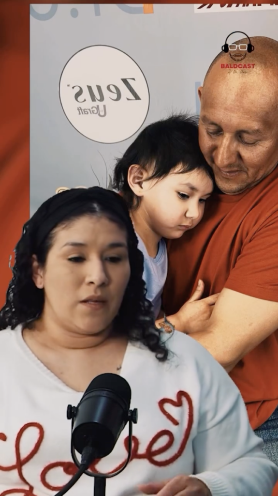 Woman speaking into a microphone with a man holding a young child in the background.