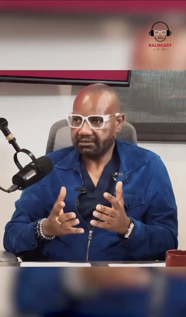 Dr. Sanusi Umar seated at a podcast table wearing white glasses and a blue jacket, speaking into a microphone.
