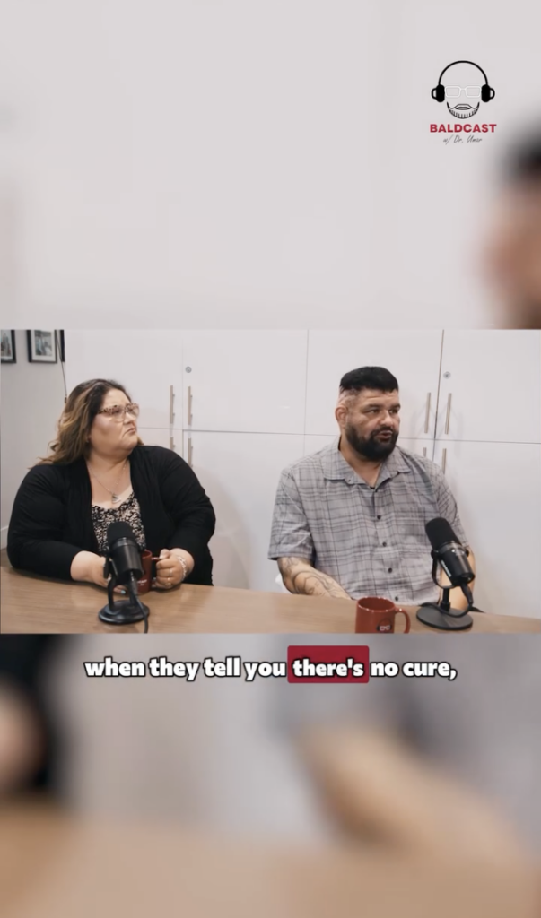 John and a woman seated at a table with microphones during a Baldcast interview.