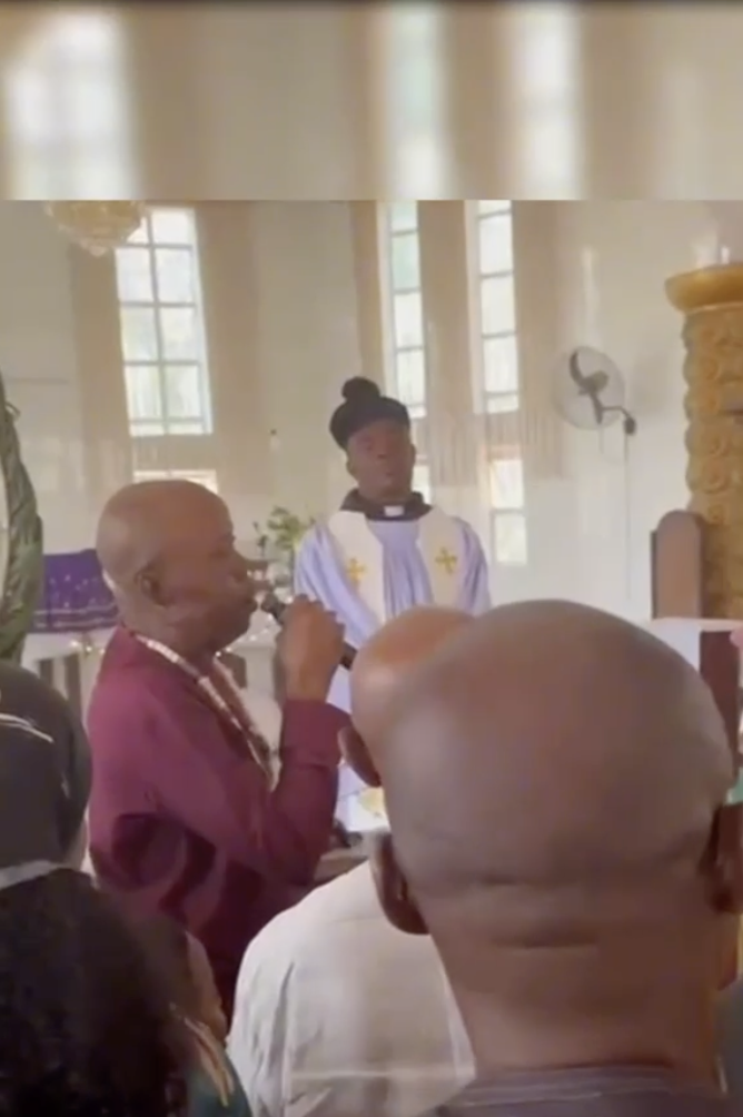 Man speaking into a microphone inside a church with a clergy member and congregants visible.