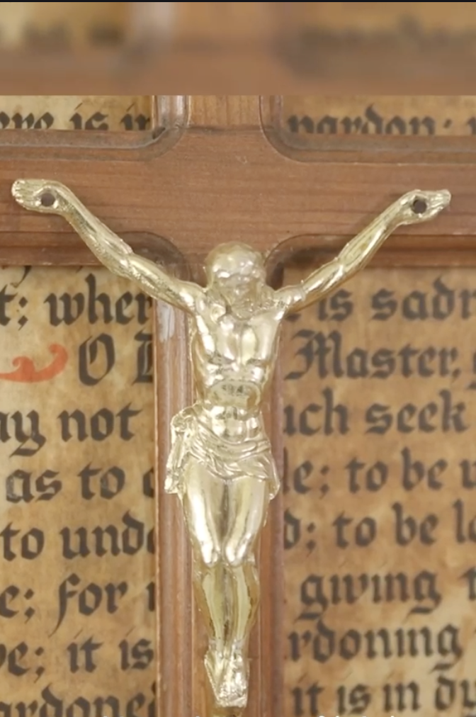 Gold-toned crucifix on a wooden cross with printed religious text in the background.