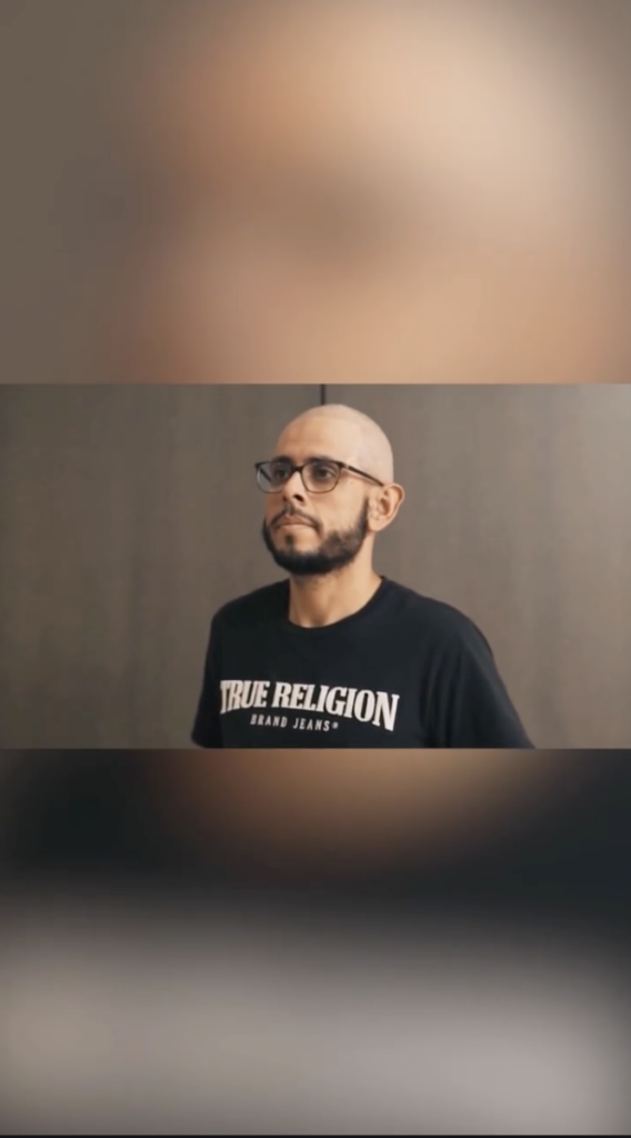 Bald man wearing glasses and a black T-shirt standing against a plain wall.