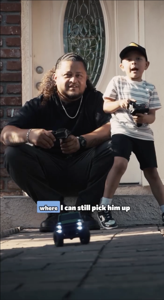 Man and young boy sitting together outside a home while holding remote controls, with a toy vehicle in the foreground.