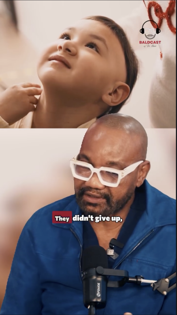 Young child looking upward above Dr. Umar seated at a microphone during a Baldcast interview.