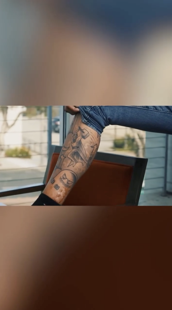 A tattooed lower leg resting against a chair, with several illustrated figures and shapes visible.