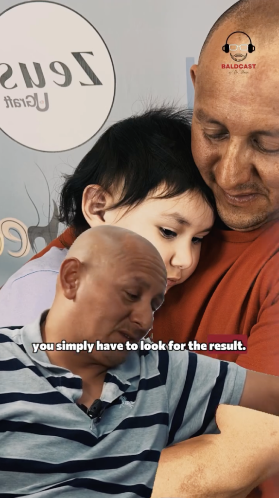 A man in a striped polo shirt speaking, with a photo of an adult holding a child behind him.