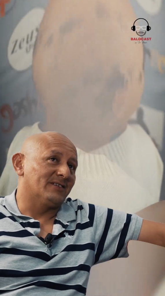 A man in a gray and navy striped polo shirt seated and speaking, with a large faded portrait behind him.