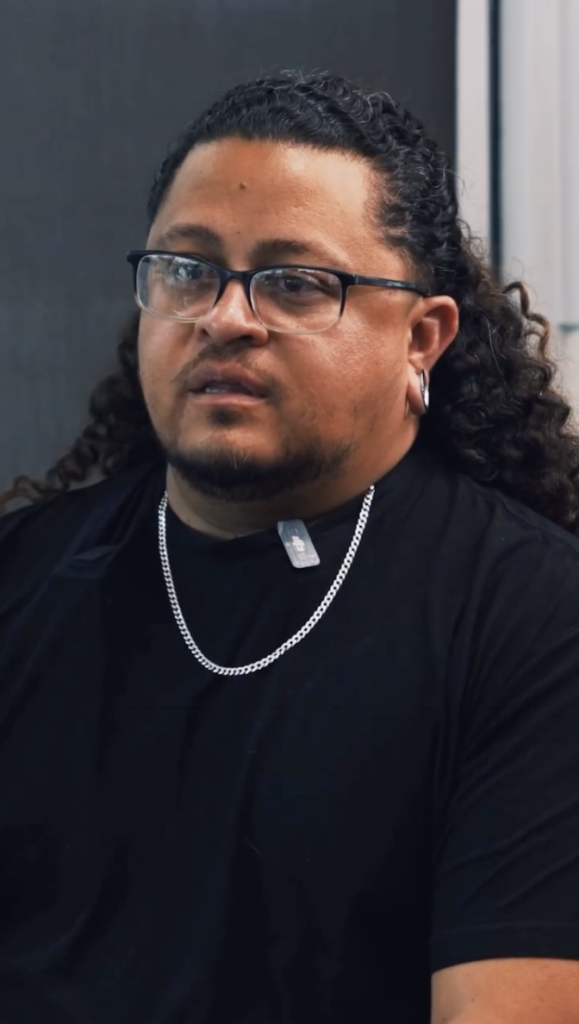 Man with glasses, long curly hair, a black shirt and a silver chain speaking while seated.