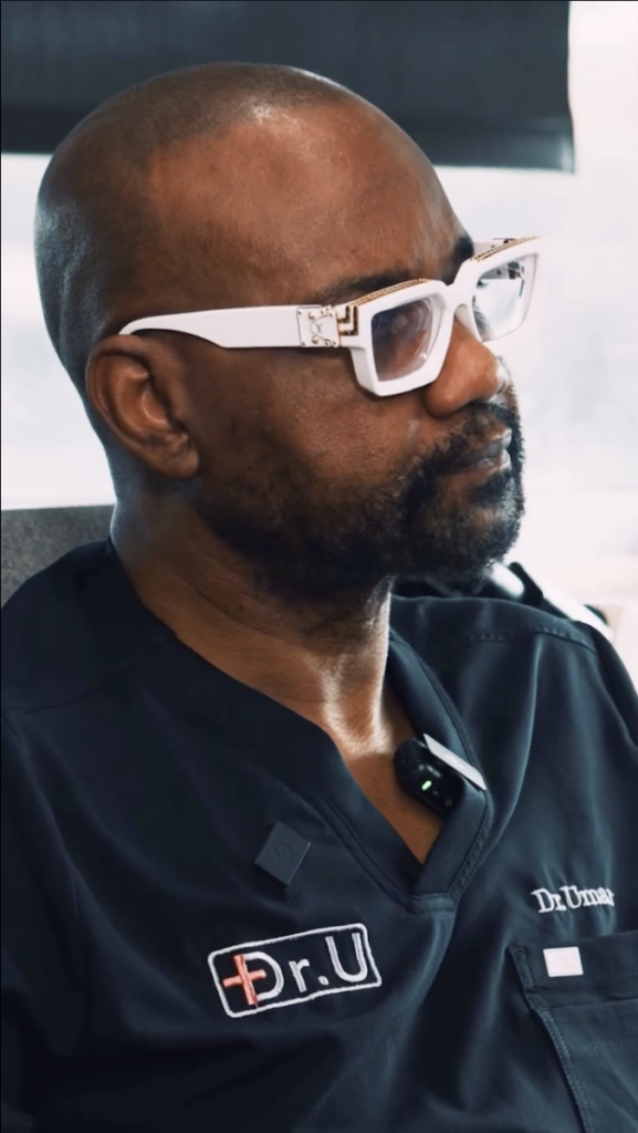 Dr. Umar in white-framed glasses and dark medical scrubs, seated and looking toward the speaker.