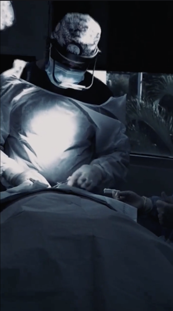 Masked clinical staff wearing surgical gowns working around a draped patient in an operating room.