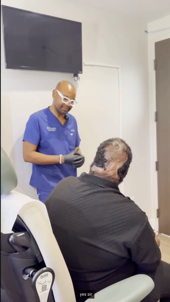 Clinician in blue scrubs and white glasses standing beside a seated man, with the back of the man’s scalp visible.