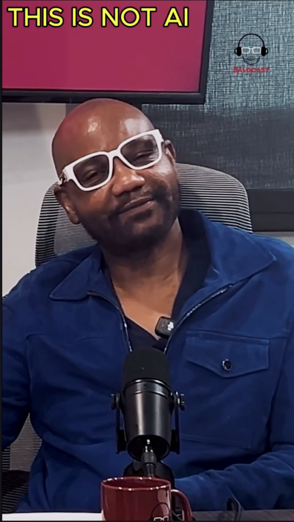 Man wearing white-framed glasses and a blue jacket speaking at a table microphone.