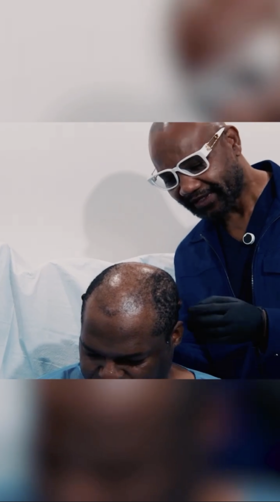 Dr. Umar, wearing white glasses and dark gloves, stands beside a seated man and looks at the top of his scalp.