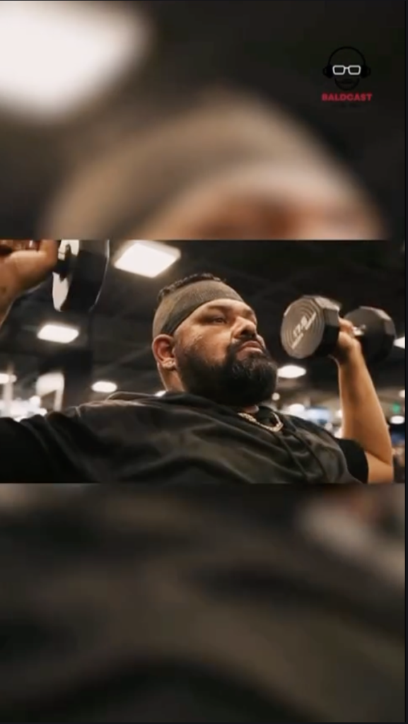 Man wearing a black shirt and headband lifting dumbbells inside a gym.