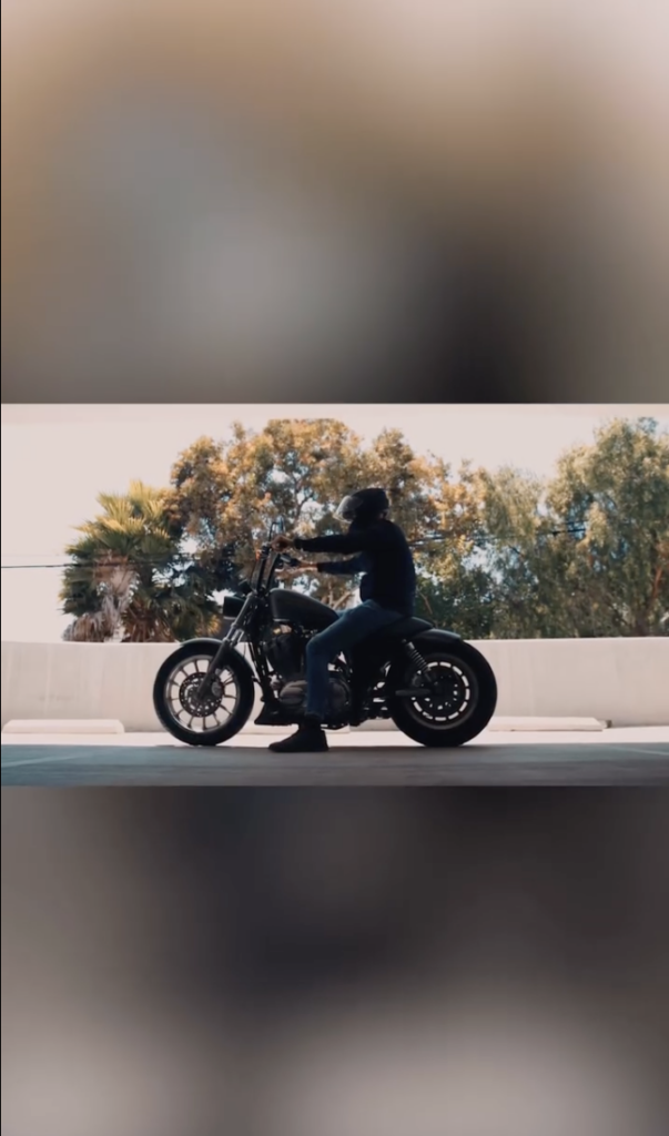 A person in dark clothing and a helmet seated on a black motorcycle outdoors with trees in the background.
