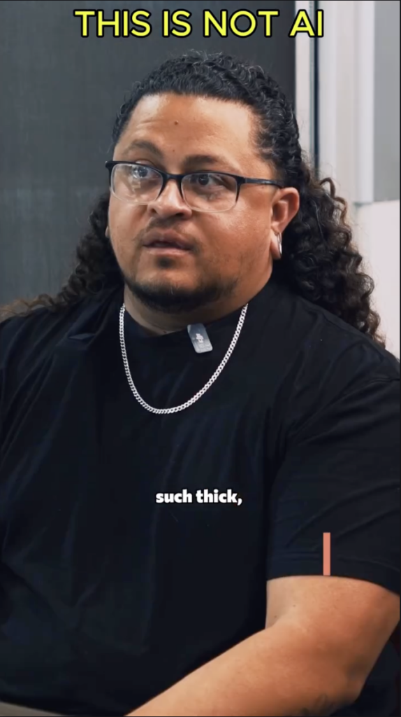 Man with long curly hair, glasses, and a black shirt seated during an interview, with the words “THIS IS NOT AI” above him.