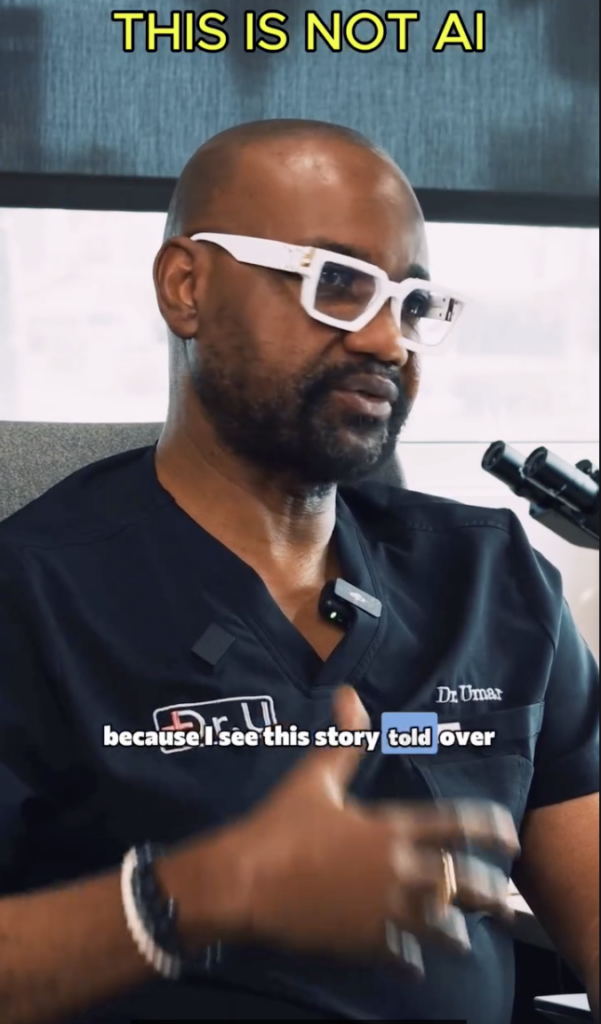 Dr. Umar wearing white glasses and dark medical scrubs, speaking beside a microphone during an interview.
