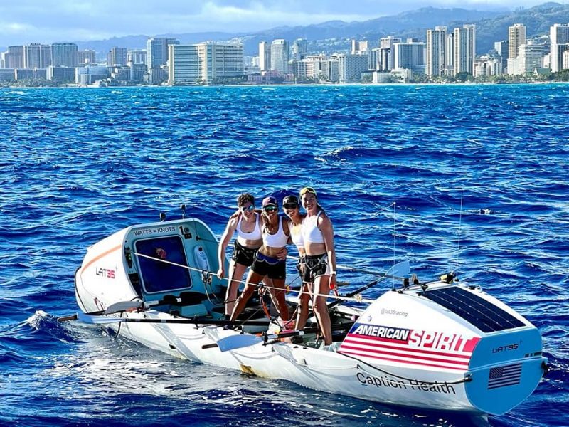 The four women rowers stand in their boat near the finish