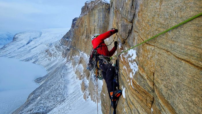 Climber maneuvres on the vertical rock wall, Greenland's ice in background.