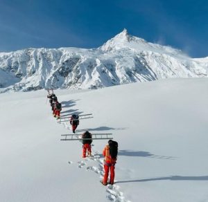 Four climbers walk in line on a flat field of fresh snow, Manaslu in front of them.