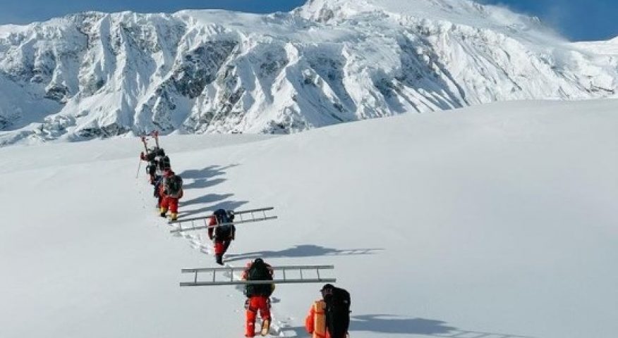 Four climbers walk in line on a flat field of fresh snow, Manaslu in front of them.