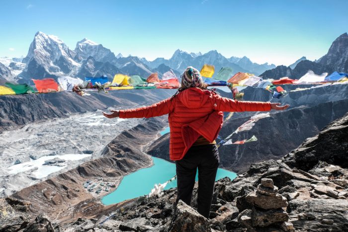 A woman opens her arms from a high place, a stunning mountain and blue lake landscape at her feet.
