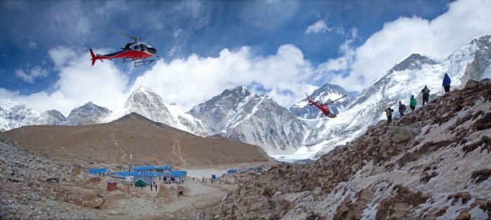 Two red helicopters, one behind the other, fly above glacial morrain terrain and cloud-capped mountains.