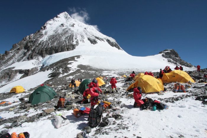 The barren South Col, snow and wind-swept rocky terrain, with some tents and climbers in the area, and Everest's summit in background.