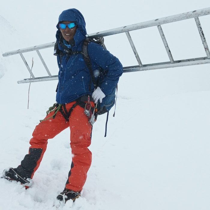 A sherpa stands with a long aluminium ladder crossed and tied to his backpack.