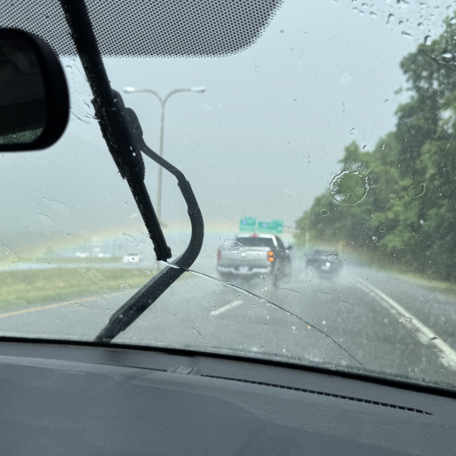 A rainbow, slightly out of focus, out of the front window of a car. The wiper is visible.