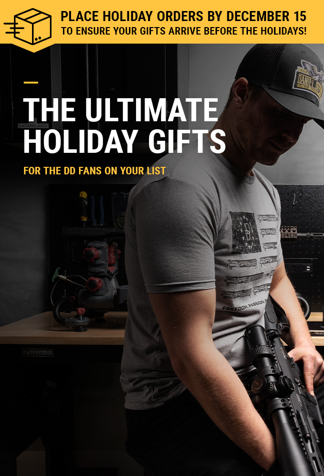 Man at work bench, wearing a black hat and gray t-shirt, holding a rifle..