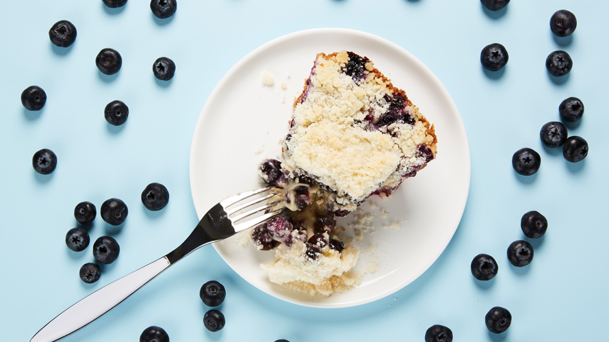 Creamy blueberry pie with sweet streusel topping served on a white plate surrounded by blueberries.