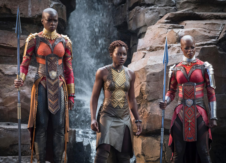 Three warrior women from Marvel's The Black Panther stand side by side. Two in bright red body armour and one in the middle wearing gold body armour.
