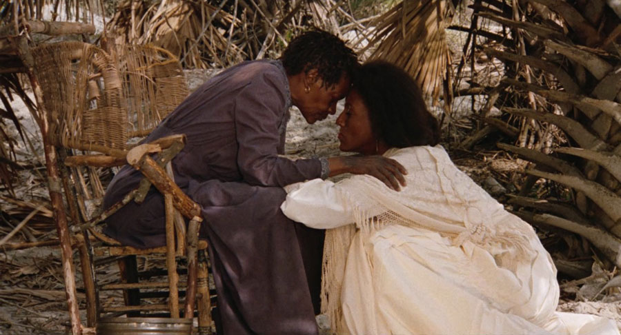 A young woman crouches in front of an older woman who is seated and grasps her legs as they press their foreheads together.