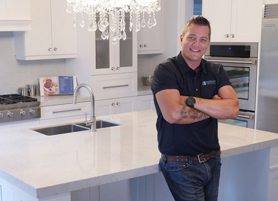 Contractor and TV personality Damon Bennett leans against the island of a kitchen featuring white cabinetry and stainless appliances.