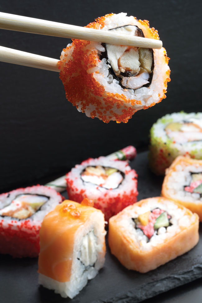 A pair of chopsticks holding a sushi roll over a plate of an assortment of sushi.