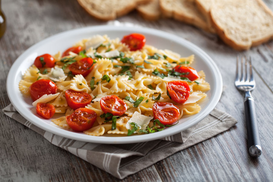A bowl of bow tie pasta with sun dried tomatoes on a wooden table with a fork and bread displayed next to it.