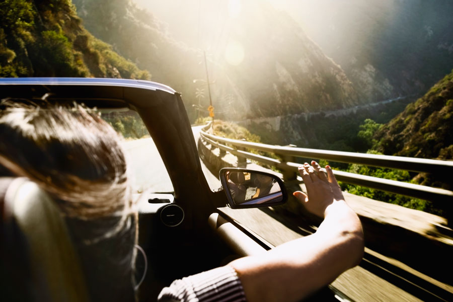 A woman driving in a convertible extending her hand out of the side of the car. Mountains can be seen surrounding the road.