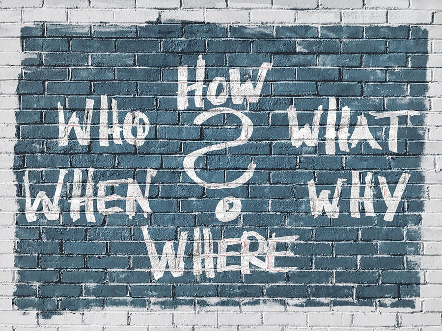 A question mark surrounded by the words how, who, what, when, why and where written on a brick wall.