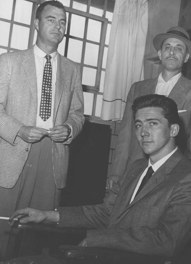 End of a 17-Day Spending Spree Cigarette in his hand, Boyne Lester Johnston sits in the office of the Denver jail building after telling of his Canadian bank theft and the spending spree that followed. Standing behind chair are Denver Detectives Clyde Villano (left) and Joe Talty.