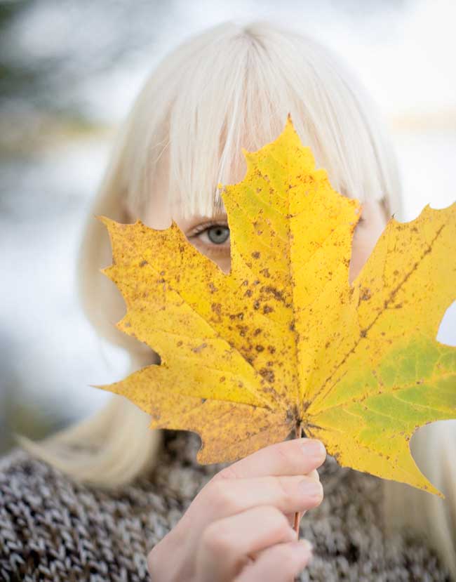 A woman holding a maple leaf in front of her face with one eye peaking out from one of the corners.