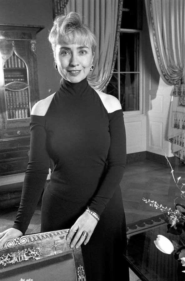 Hillary Clinton in a black off-the-shoulder dress sitting on a desk smiling.