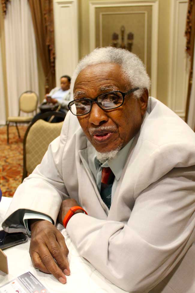 A man with grey hair leaning on a desk wearing thick black framed glasses.