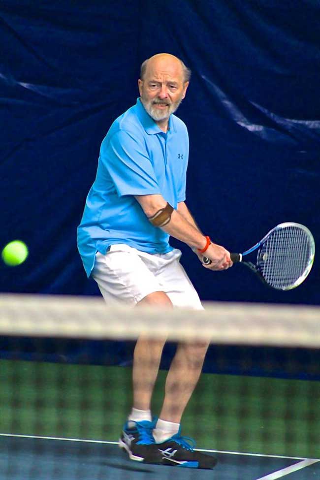 Older balding man prepares to take a swing at a tennis ball with his tennis racket.