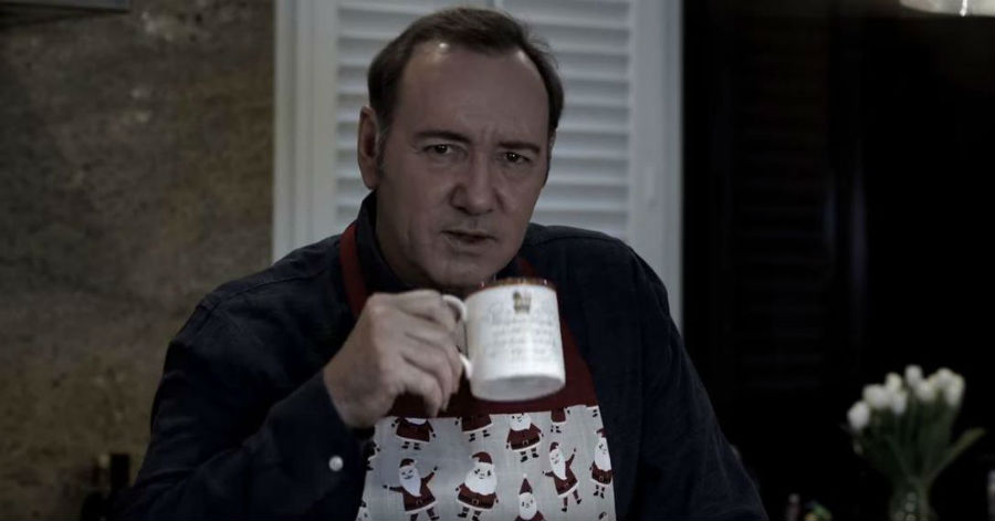 Kevin Spacey wearing a snowman print holiday sweater and holding a coffee cup.