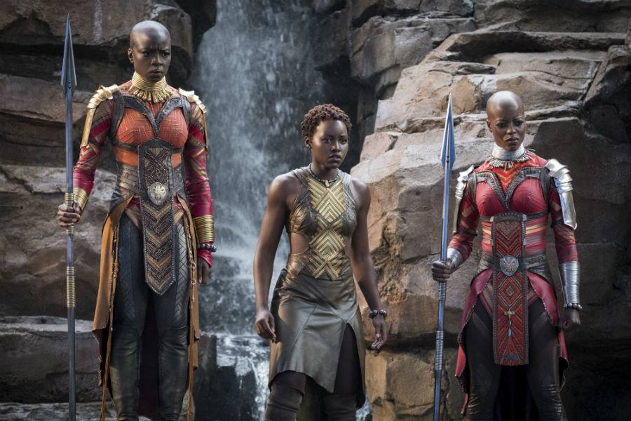 Three Wakanda warrior women stand in front of a waterfall, each wearing futuristic armour with a tribal element to it as well.