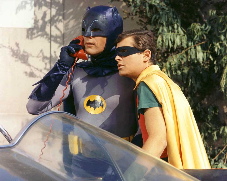 Batman and Robin stand by the batmobile as batman takes a call on the batphone.