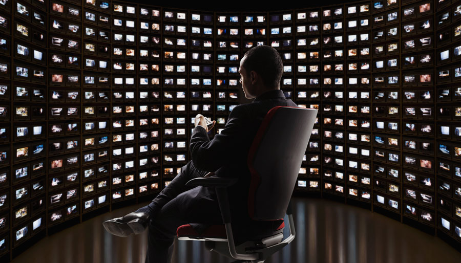 A Man sits in an office chair in front of a wall of TV screens