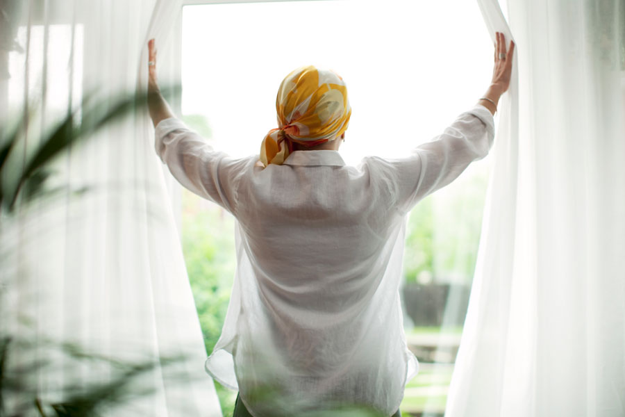 A woman wearing a bandana opens sheer drapes and looks out the window.